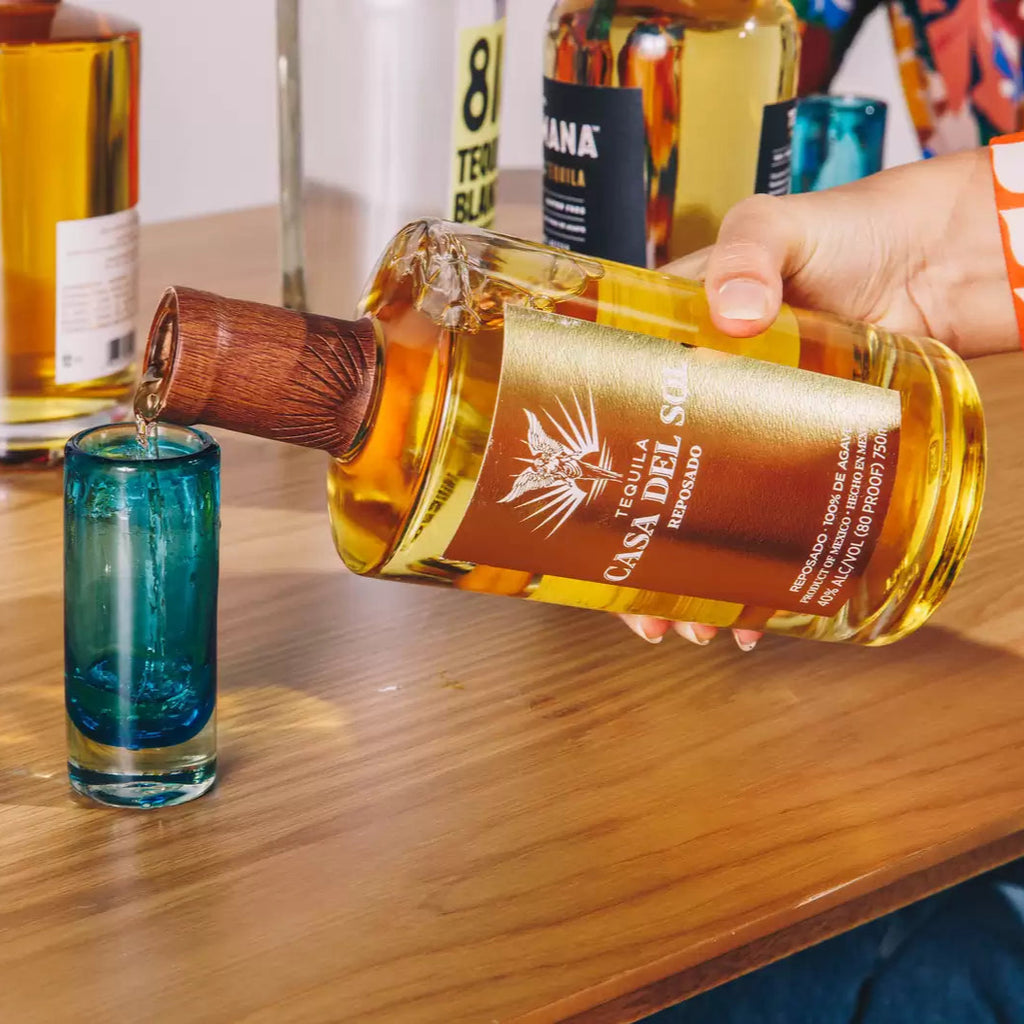 Person pouring Casa Del Sol tequila into a shot glass on a wooden table.