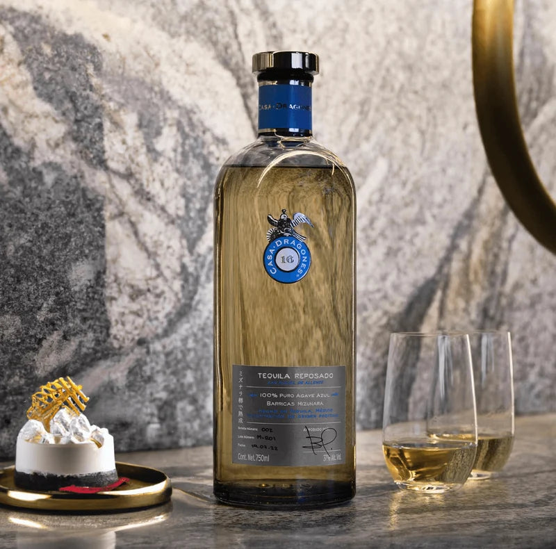 Load image into Gallery viewer, A bottle of Casa Dragones Reposado Mizunara Tequila is displayed on a marble surface next to a glass of the aged tequila and a small dessert, set against a grey marble wall background.