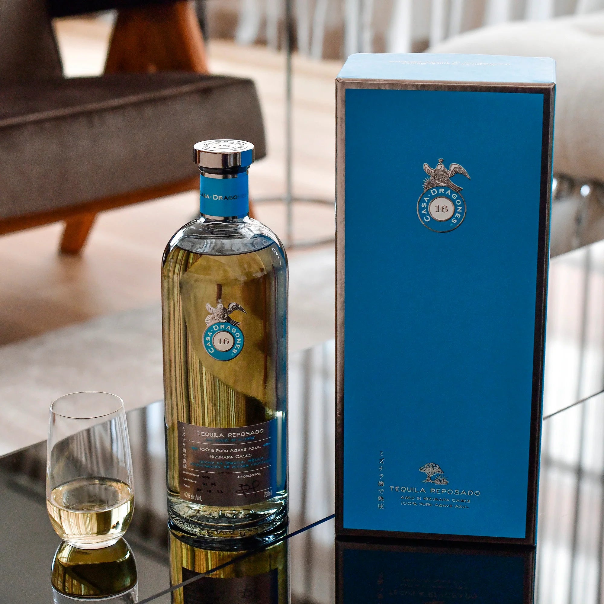 Load image into Gallery viewer, A bottle of Casa Dragones Reposado Mizunara Tequila stands on a glass table beside a matching blue presentation box and a small filled glass, with a softly blurred indoor room in the background.