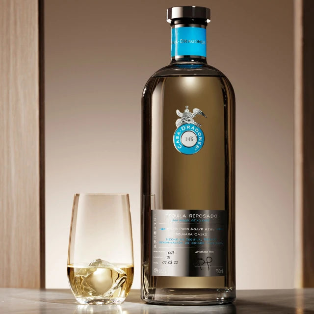 Load image into Gallery viewer, A bottle of Casa Dragones Reposado Mizunara Tequila with a clear glass of the golden liquid beside it, set on a reflective dark surface against a warm beige background.
