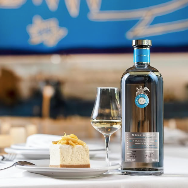 Load image into Gallery viewer, A bottle of Casa Dragones Reposado Mizunara Tequila on a white tablecloth next to a wine glass and a slice of cheesecake, with a blurred restaurant background featuring a blue sign.