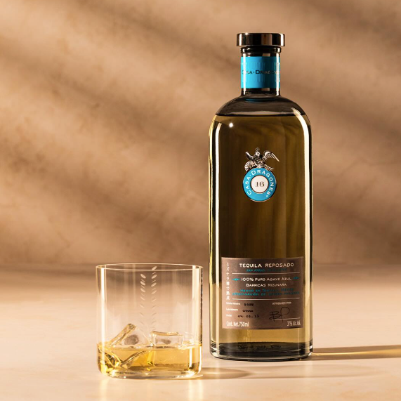 Load image into Gallery viewer, A bottle of Casa Dragones Reposado Mizunara Tequila alongside a clear rocks glass with ice, set against a warm beige background with soft diagonal shadows.