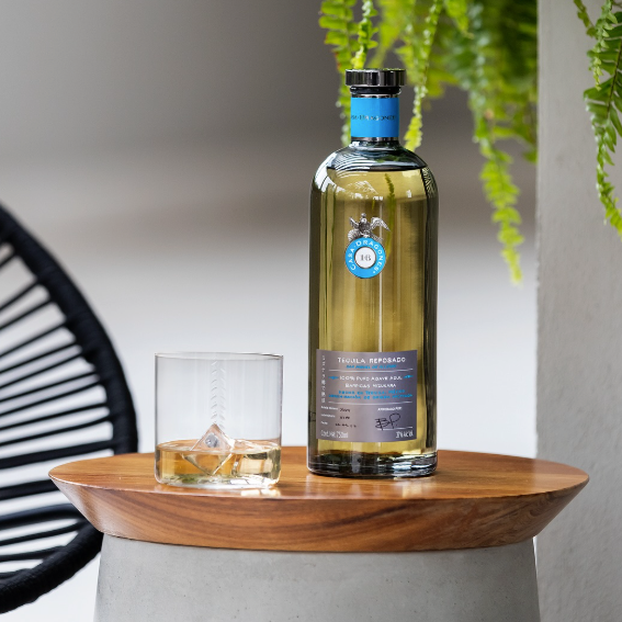 Load image into Gallery viewer, A bottle of Casa Dragones Reposado Mizunara Tequila with a matching glass on a round wooden side table, styled with a hanging green plant and a black chair in the background.