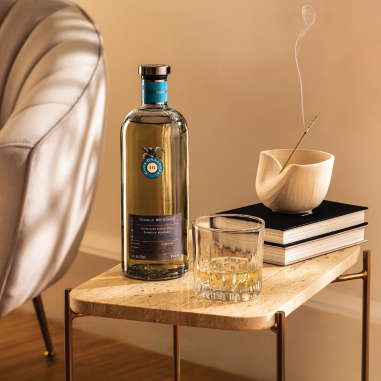 Load image into Gallery viewer, A bottle of Casa Dragones Reposado Mizunara Tequila sits on a marble-topped side table next to a filled rocks glass, stacked books, and a smoking incense holder in a warm, modern interior setting.