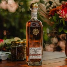 Load image into Gallery viewer, Casa Noble tequila bottle on a wooden surface with a floral background