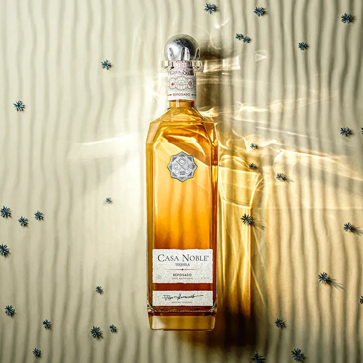 Load image into Gallery viewer, A bottle of Casa Noble Tequila Reposado standing upright on a textured beige surface with dramatic side lighting and scattered small blue star-like confetti.