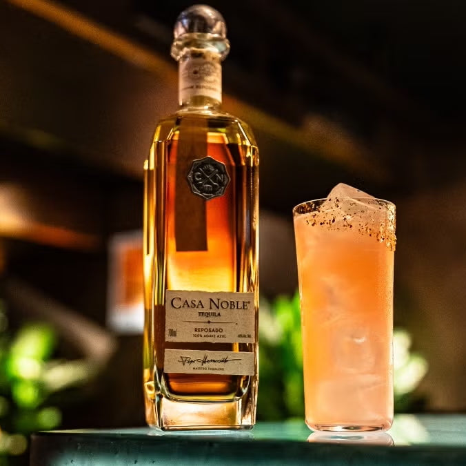Load image into Gallery viewer, Bottle of Casa Noble tequila next to a cocktail on a bar with a blurred background