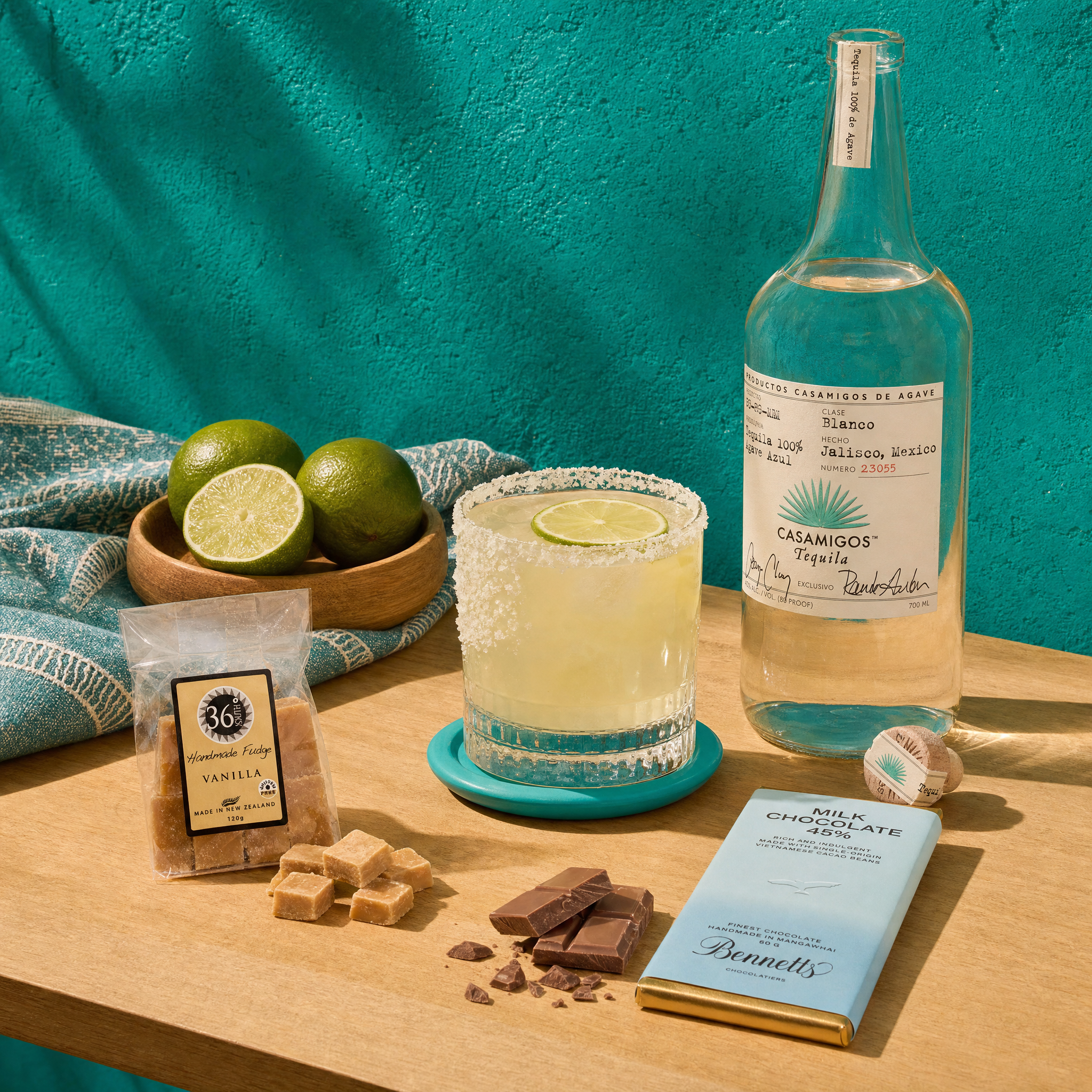 Load image into Gallery viewer, Casamigos blanco tequila bottle, margarita, limes, vanilla beans, chocolate, and a bar of chocolate on a wooden surface with a teal background.