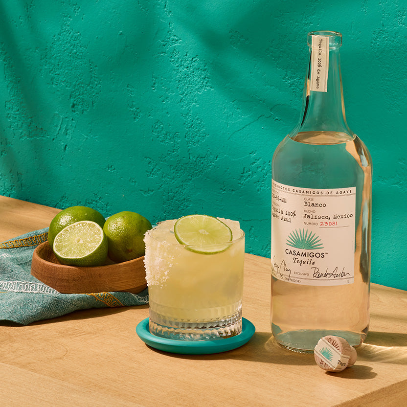 Load image into Gallery viewer, Casamigos blanco tequila bottle with a margarita and limes on a wooden surface against a teal wall.