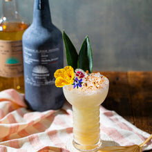 Load image into Gallery viewer, Casamigos Mezcal Cocktail with floral garnish in a glass, with bottles in the background on a checkered cloth.