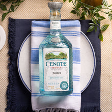 Load image into Gallery viewer, Cenote Blanco Tequila bottle on a blue and white striped towel with greenery in the background