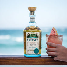 Load image into Gallery viewer, Cenote reposado tequila bottle with a cocktail on a wooden surface by the ocean.