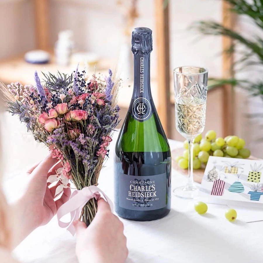 Load image into Gallery viewer, A bottle of champagne on a table with some grapes and a woman holding a bouquet of dried flowers infront of the bottle
