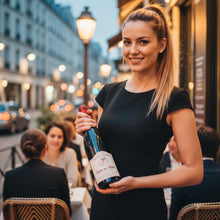 Load image into Gallery viewer, Woman holding a bottle of Charvin Cotes du Rhone wine in an outdoor setting with blurred people and lights in the background