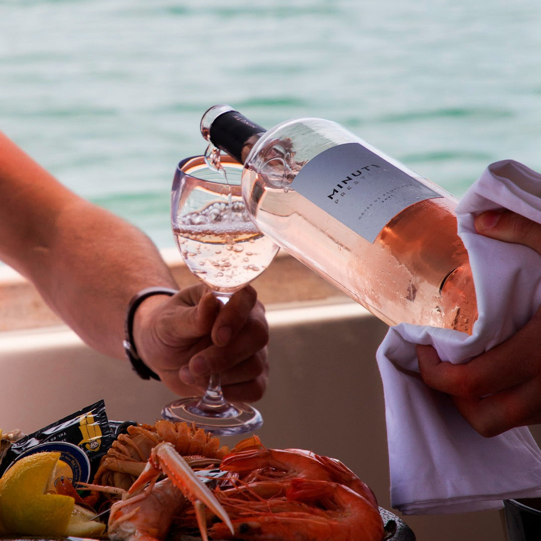 Load image into Gallery viewer, Person pouring rosé wine into a glass with a plate of prawns and lemon wedges by the sea.