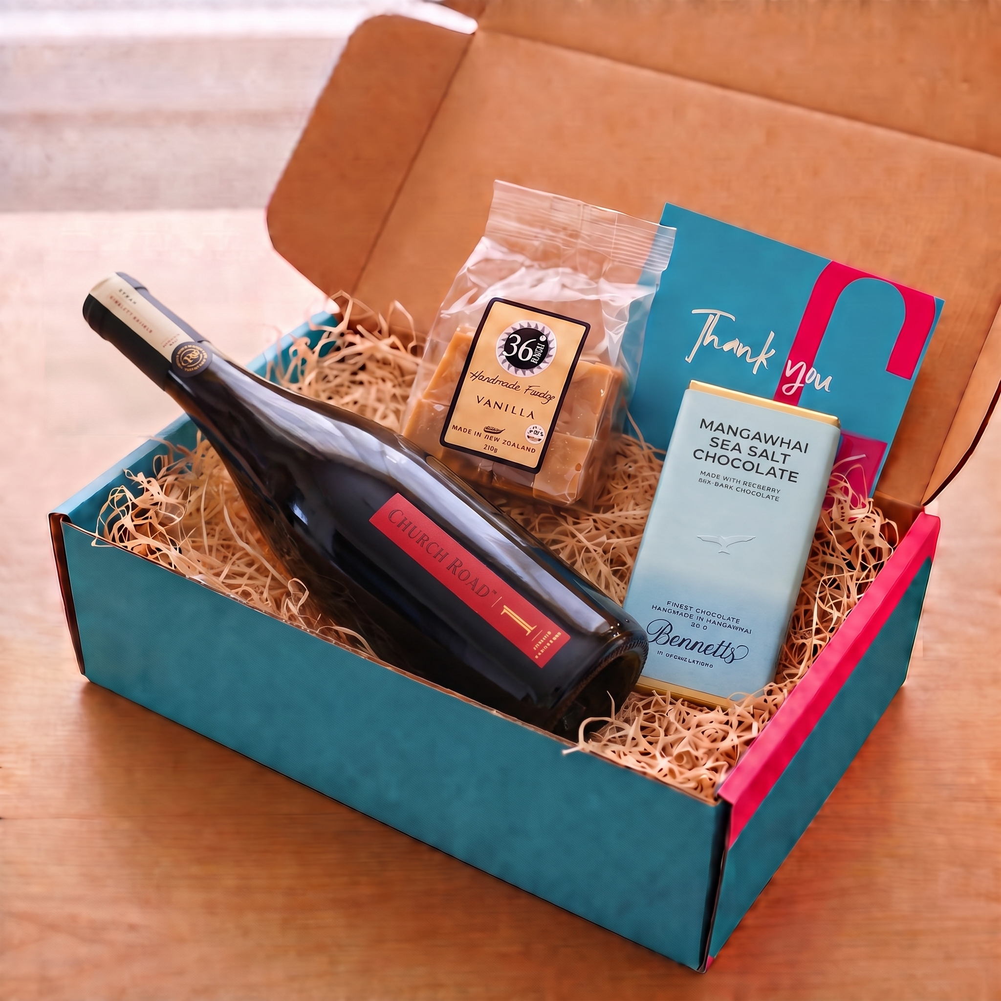 Load image into Gallery viewer, A curated gift box set on a wooden table, featuring a bottle of Church Road 1 Syrah red wine, a bag of 36 Degrees Vanilla fudge, a bar of Bennett's Mangawhai Sea Salt Chocolate, and a blue and red thank you card, all arranged in a teal and pink cardboard box with wood-wool filler.