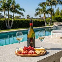 Load image into Gallery viewer, A bottle of Church Road Grand Reserve Chardonnay next to two wine glasses and a platter of meat and cheeses beside a pool surrounded by palm trees.