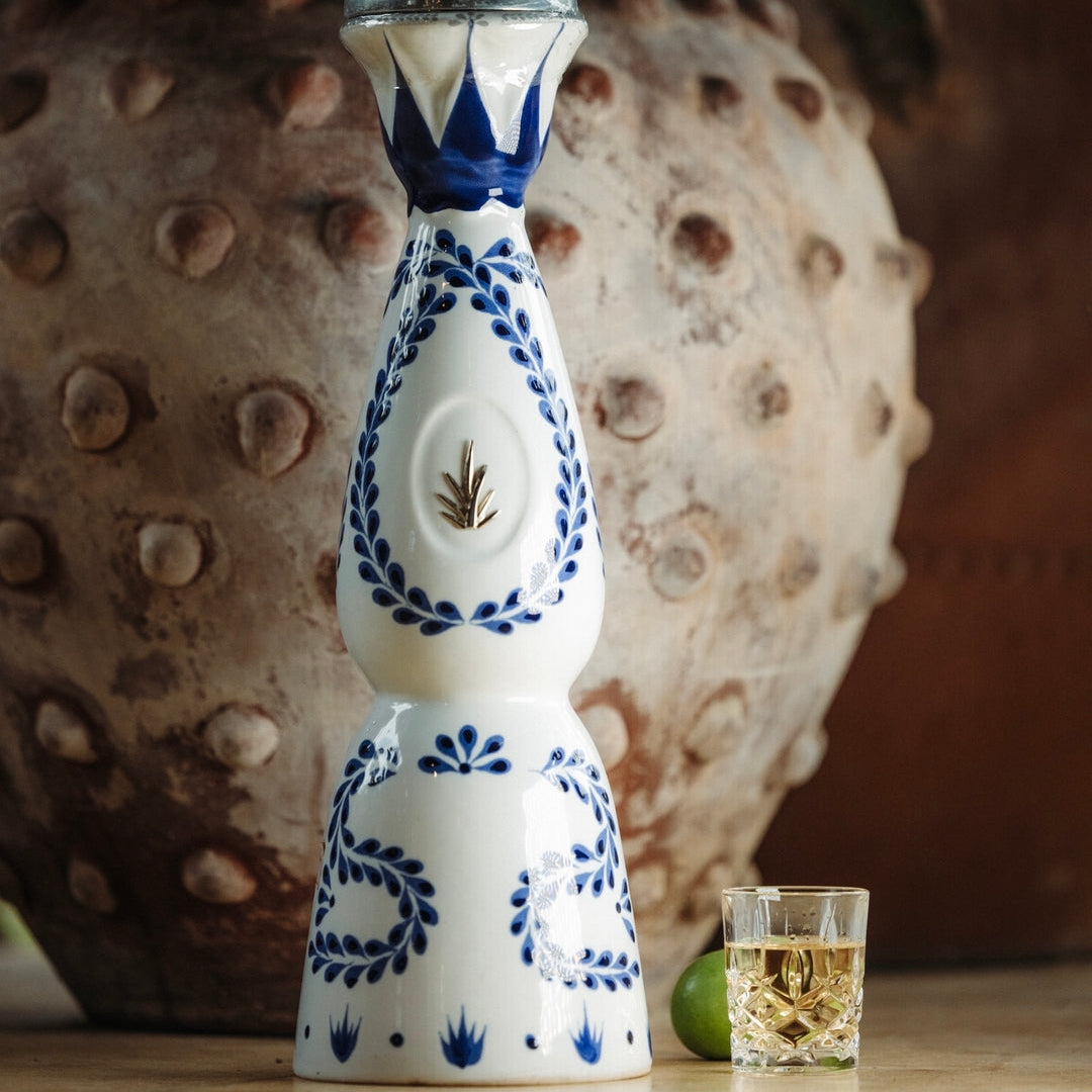 Load image into Gallery viewer, A Clase Azul Reposado Tequila ceramic bottle with a glass top on a wooden surface, with a textured vase and glasses in the background.