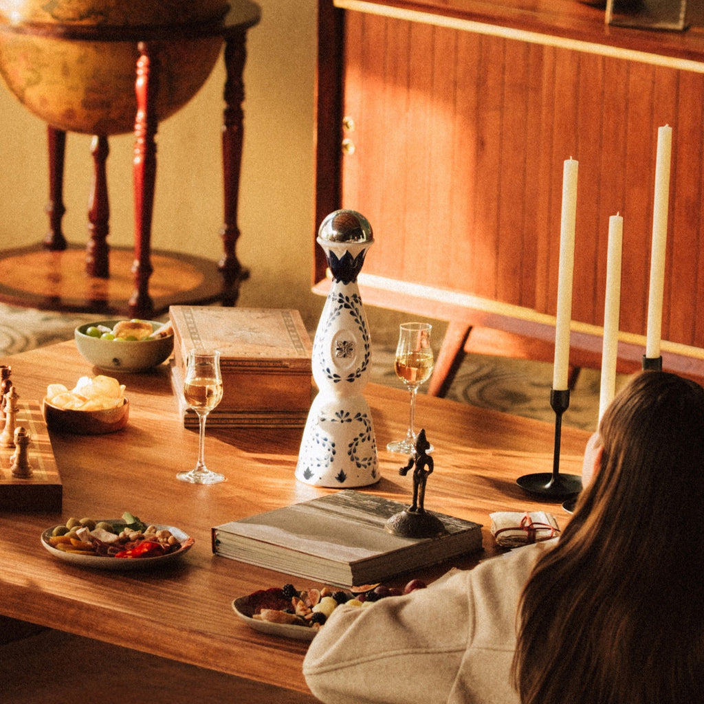 Person sitting at a wooden table with a bottle of Clase Azul Reposado Tequila and decorative items, candles, and a globe in the background.