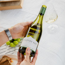 Load image into Gallery viewer, Person holding a bottle of Cloudy Bay wine with a glass and grapes on a table.