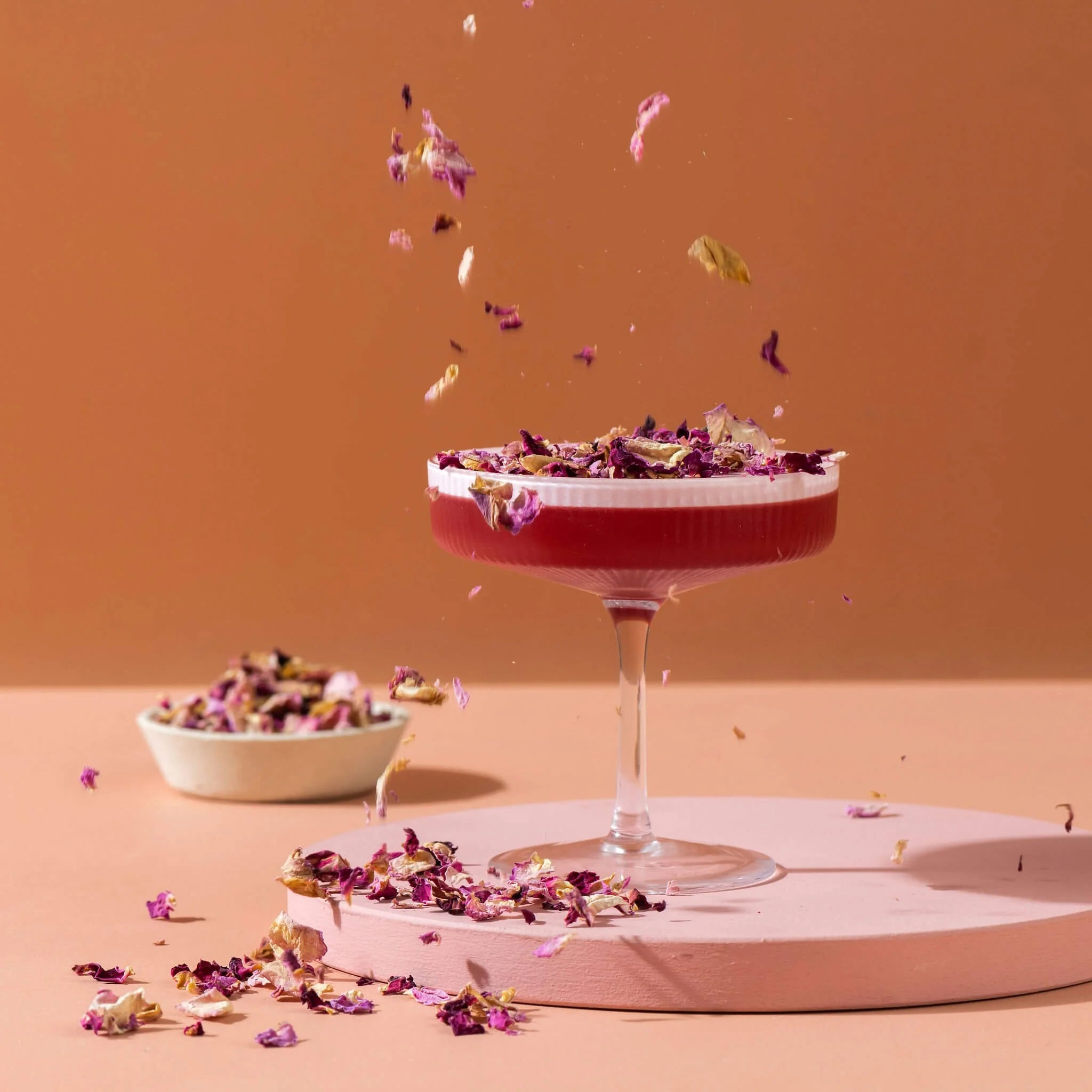 Load image into Gallery viewer, Cocktail glass with dried edible flowers on a pink platform against an orange background