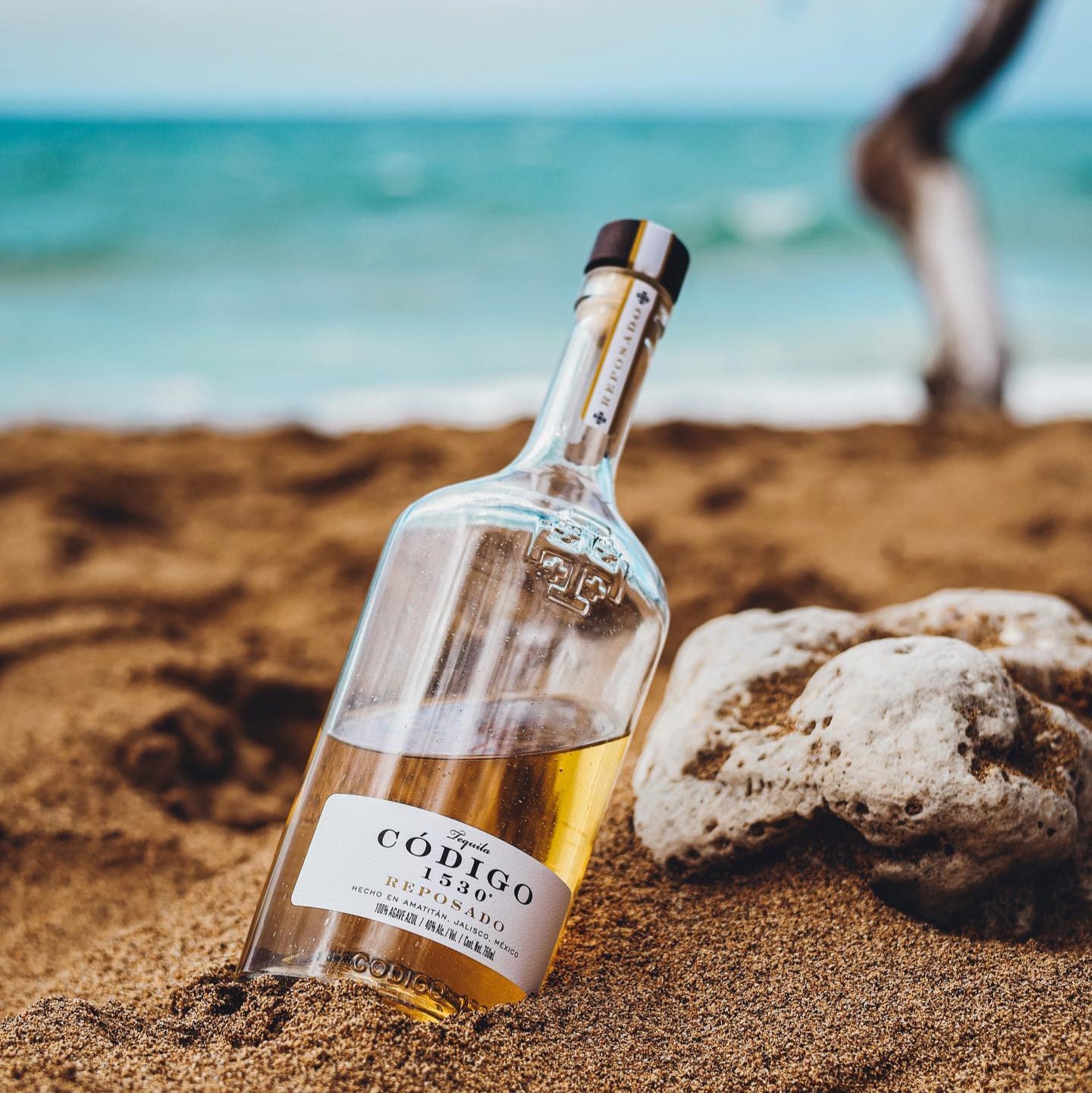 Load image into Gallery viewer, Bottle of Códico reposado tequila on a sandy beach with ocean in the background