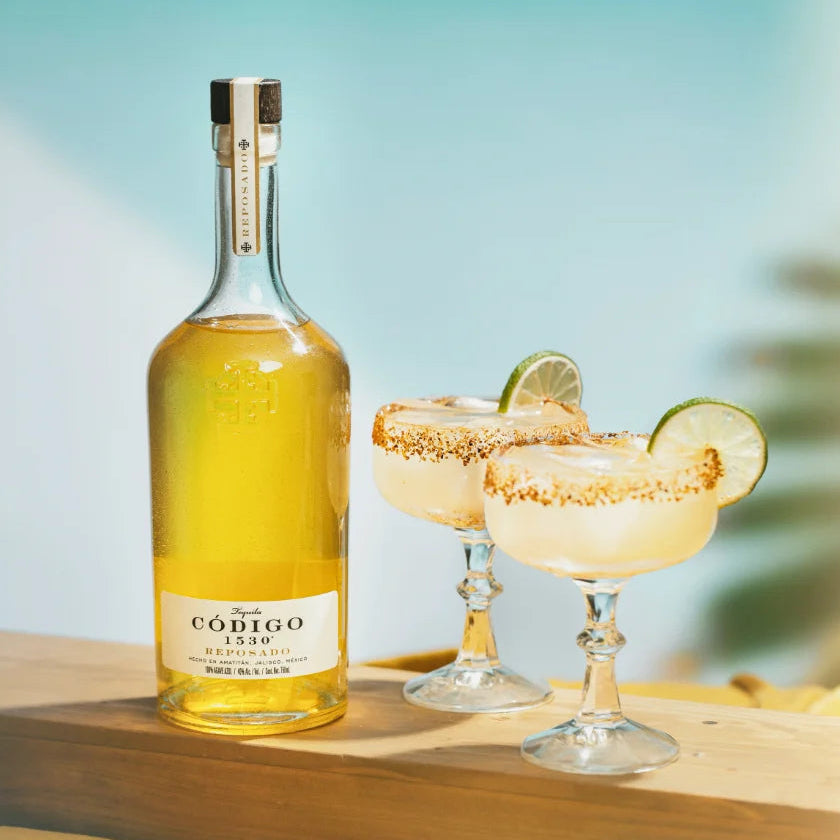 Load image into Gallery viewer, Bottle of Código 1530 reposado tequila with two margaritas on a wooden surface against a blue sky.