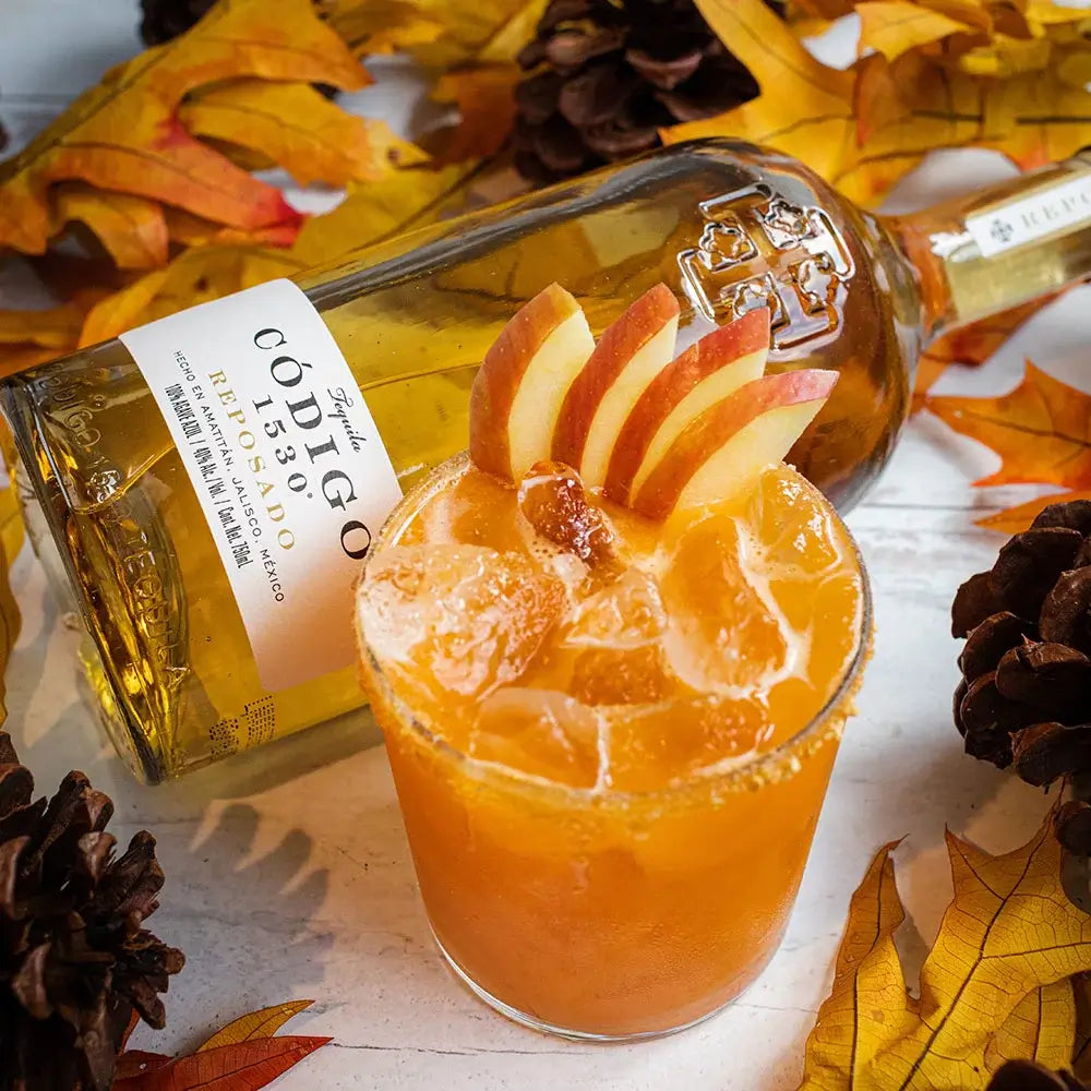 Load image into Gallery viewer, Bottle of Codigo Reposado tequila lying on its side next to a orange cocktail with apple garnish