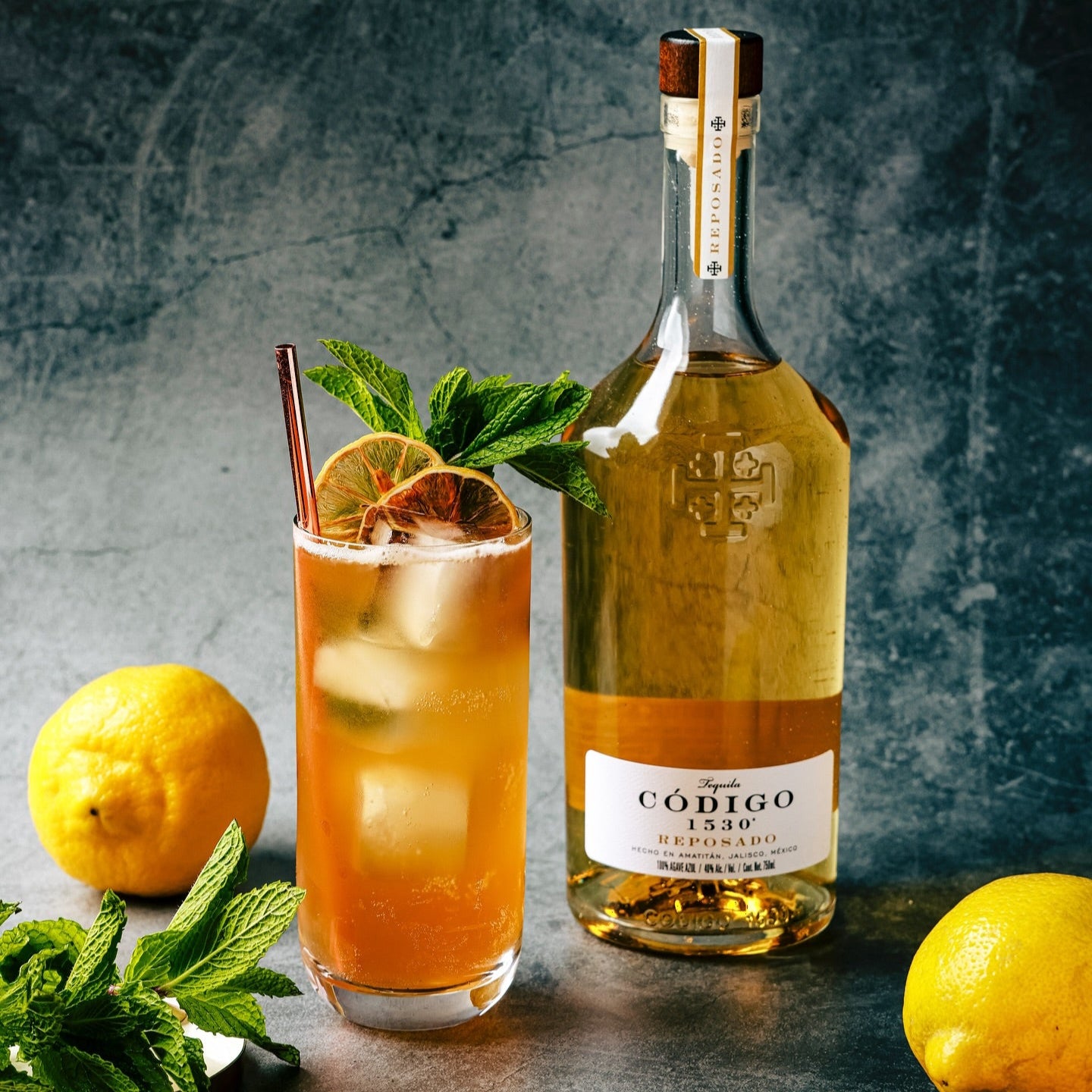 Load image into Gallery viewer, Bottle of Código reposado tequila with a cocktail garnished with lemons and mint leaves on a textured surface.