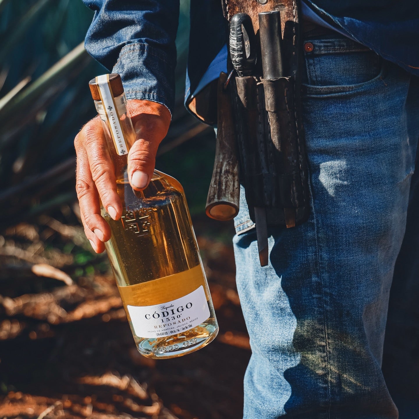 Load image into Gallery viewer, Person holding a bottle of Código reposado tequila with a rifle over their shoulder outdoors.