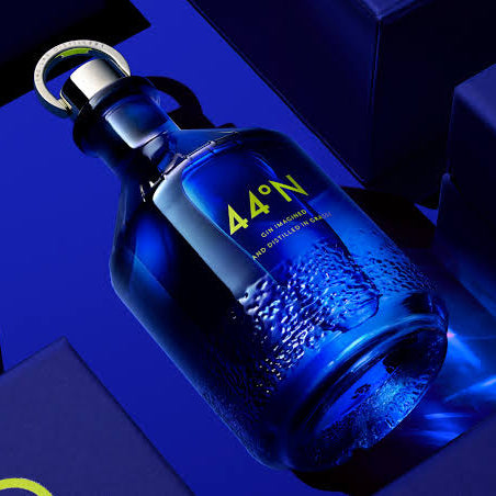 Load image into Gallery viewer, A blue glass bottle of Comte de Grasse 44°N gin, shown at an angle with condensation droplets, surrounded by dark blue packaging boxes with yellow accents.