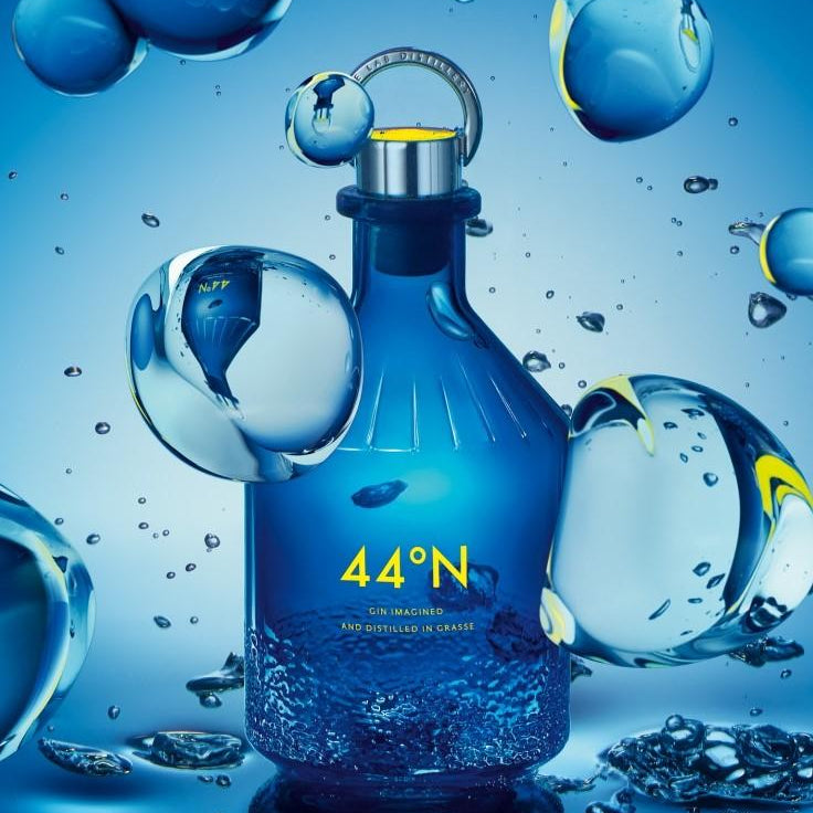 Load image into Gallery viewer, A blue glass bottle of Comte de Grasse  44°N gin with a silver cap and yellow logo, surrounded by floating water droplets and ice spheres on a blue background.