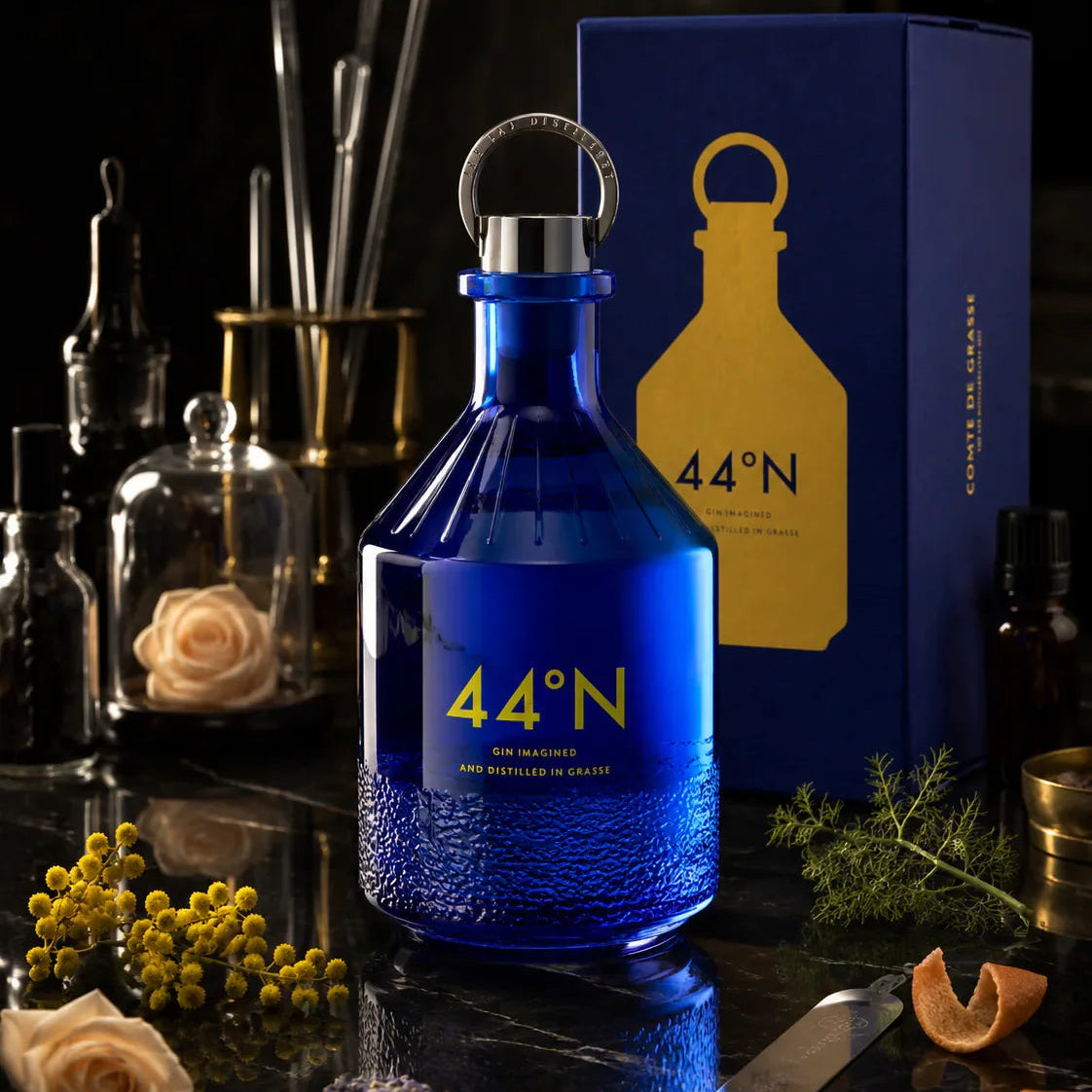 Load image into Gallery viewer, A blue bottle of Comte de Grasse 44°N gin with a silver ring stopper, displayed next to its dark blue packaging box with a gold logo, surrounded by botanical and bar-themed props on a dark reflective surface.