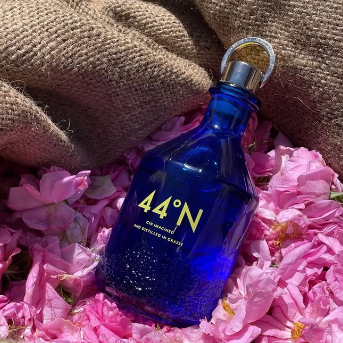 Load image into Gallery viewer, A cobalt blue glass bottle of Comte de Grasse 44 degrees N Gin with gold lettering, resting on a bed of pink flowers with a brown burlap fabric background.