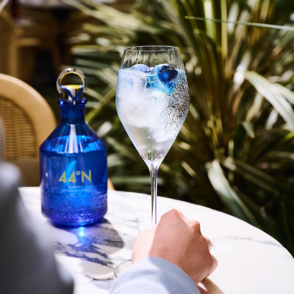 Cocktail in a glass with ice and blueberries next to a blue bottle of Comte-de-Grasse-44-degrees-N-Gin on a table outdoors.