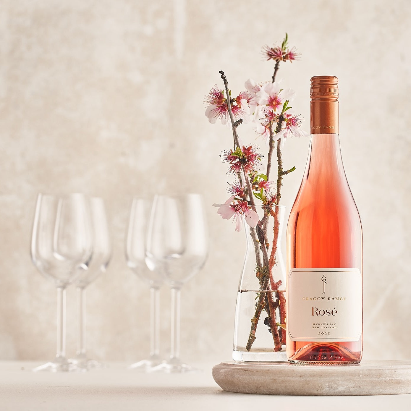 Load image into Gallery viewer, A bottle of Craggy Range Hawke's Bay Rosé on a white table and a vase of blossom and wineglasses in the background