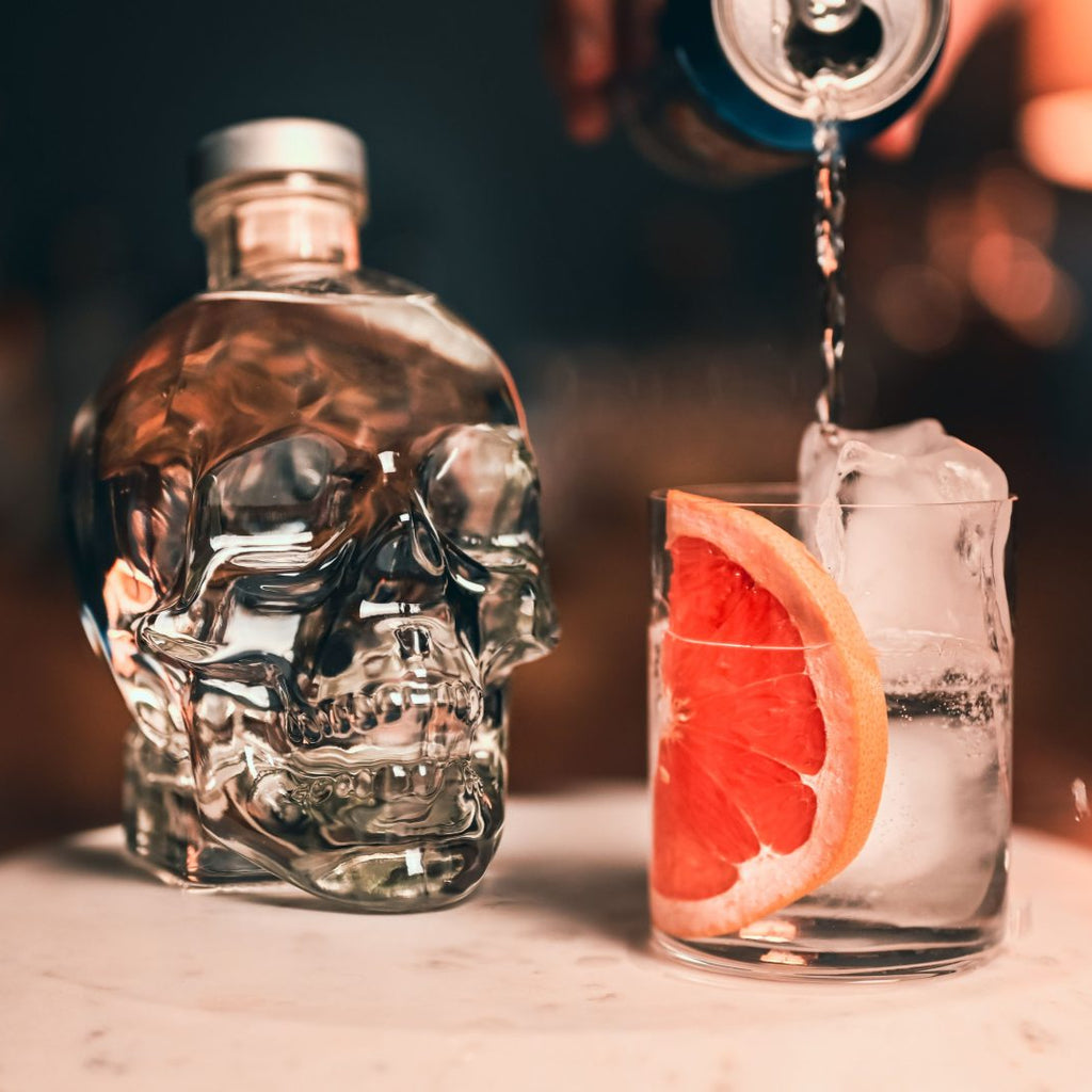 Crystal Head bottle with a cocktail being poured into a glass with a grapefruit slice on a bar.