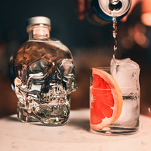 Load image into Gallery viewer, Crystal Head bottle with a cocktail being poured into a glass with a grapefruit slice on a bar.