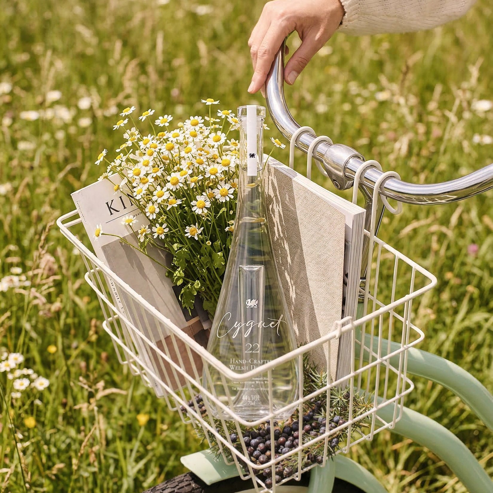 Load image into Gallery viewer, A stunning clear glass bottle of Cygnet 22 Welsh Dry Gin is placed in a white wire bicycle basket on a mint green bike, alongside daisies and magazines, with a person's hand resting on the handlebar in a grassy field.