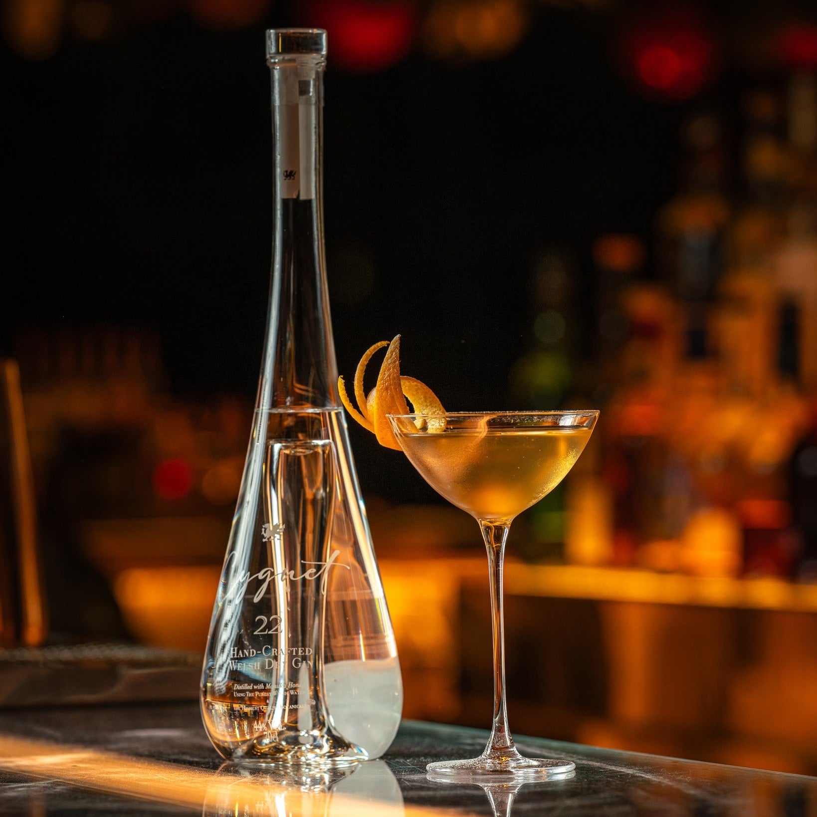 Load image into Gallery viewer, A clear glass bottle of Cygnet 22 Welsh Dry Gin and matching coupe-style cocktail glass filled with an amber drink, set on a bar counter with a blurred bar background and warm lighting.