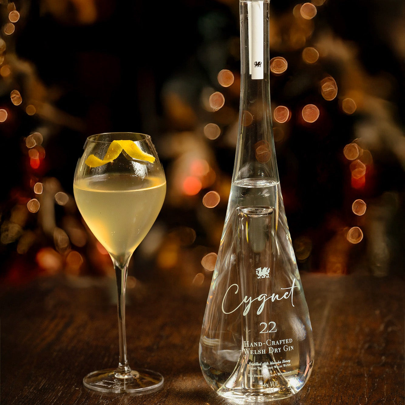Load image into Gallery viewer, A stunning clear glass bottle of Cygnet 22 Welsh Dry Gin stands on a dark wooden table beside a stemmed glass filled with pale golden liquid and a lemon twist, against a warm blurred holiday background with bokeh lights.