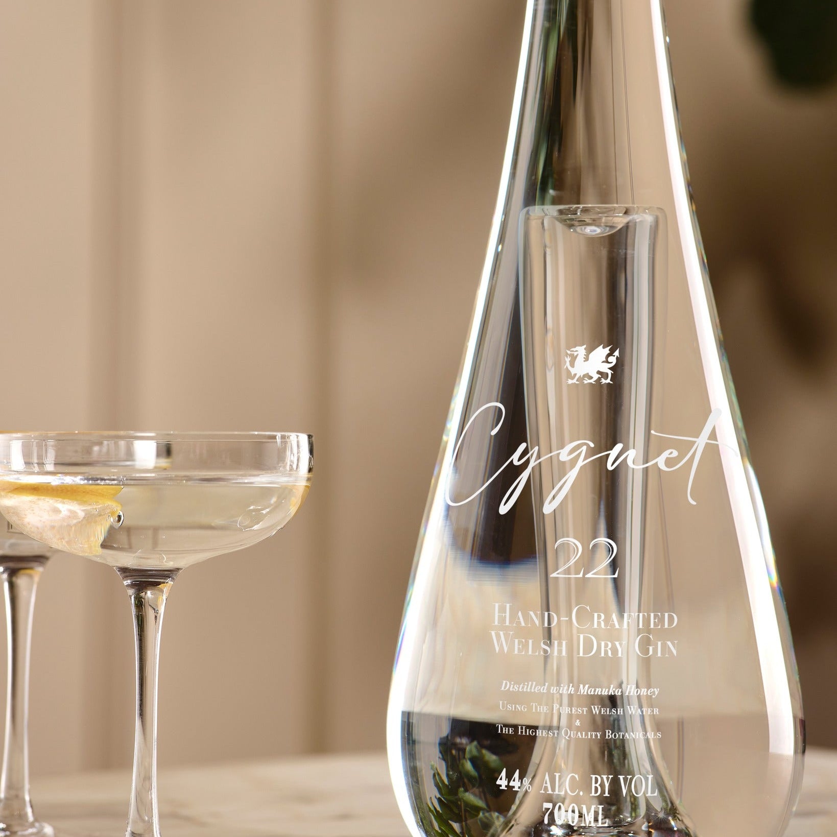Load image into Gallery viewer, A beautiful clear glass bottle of Cygnet 22 Welsh Dry Gin beside a coupe glass with a pale gin and a lemon slice, set against a softly blurred warm beige background.