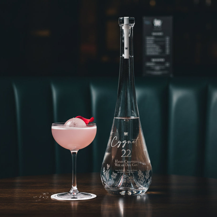 Load image into Gallery viewer, A clear glass bottle of Cygnet 22 Welsh Dry Gin stands on a dark wooden table beside a coupe glass filled with pink cocktail drink and a red garnish, set in a dimly lit bar or restaurant interior with green leather seating in the background.