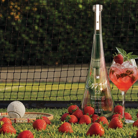 Load image into Gallery viewer, A tall clear bottle of Cygnet 22 Welsh Dry Gin stands on grass tennis court outdoors, surrounded by fresh strawberries, with a pink cocktail glass and a white ball near it. A black net and blurred greenery fill the background.