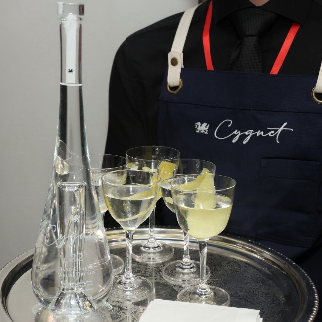 Load image into Gallery viewer, A bartender in a navy blue apron with 'Cygnet' embroidered on it holds a silver tray with a clear glass bottle of Cygnet 22 Welsh Dry Gin, three filled cocktail glasses with lemon slices, and folded white napkins.