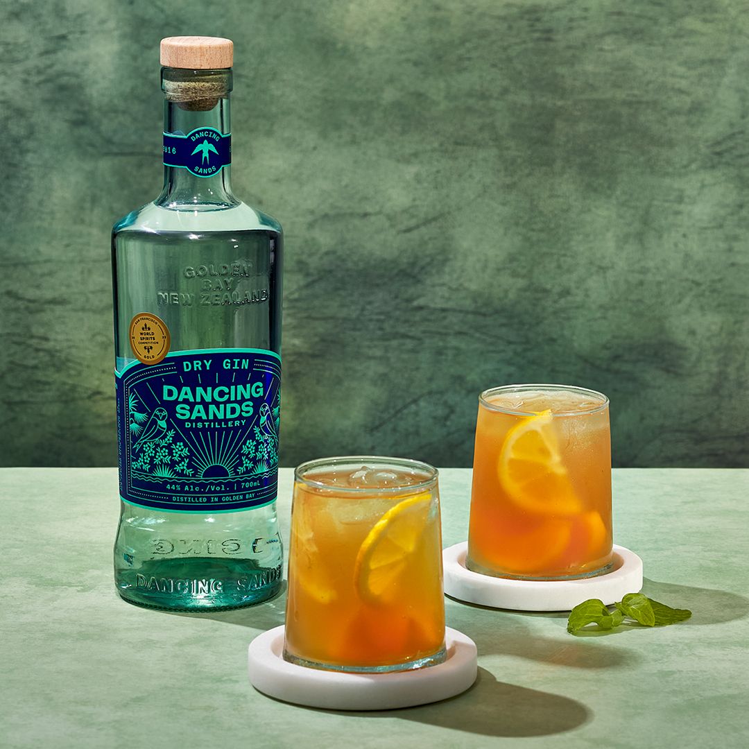 Load image into Gallery viewer, Bottle of Dancing Sands Dry Gin with two glasses of orange cocktails on a green background
