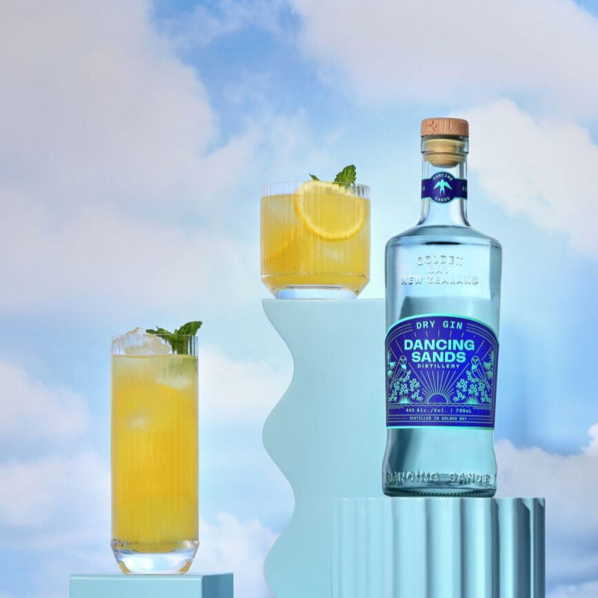 Load image into Gallery viewer, Bottle of Dancing Sands gin with two glasses of lemonade against a cloudy sky background