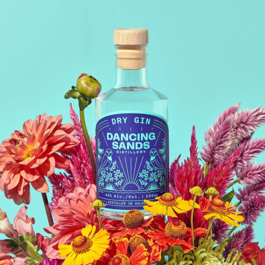 Bottle of Dancing Sands Dry Gin surrounded by colorful flowers on a bright blue background