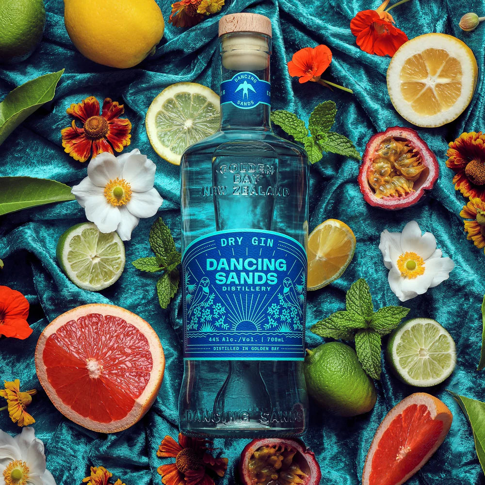 Load image into Gallery viewer, Bottle of Dancing Sands Dry Gin surrounded by fruits and flowers on a blue fabric background