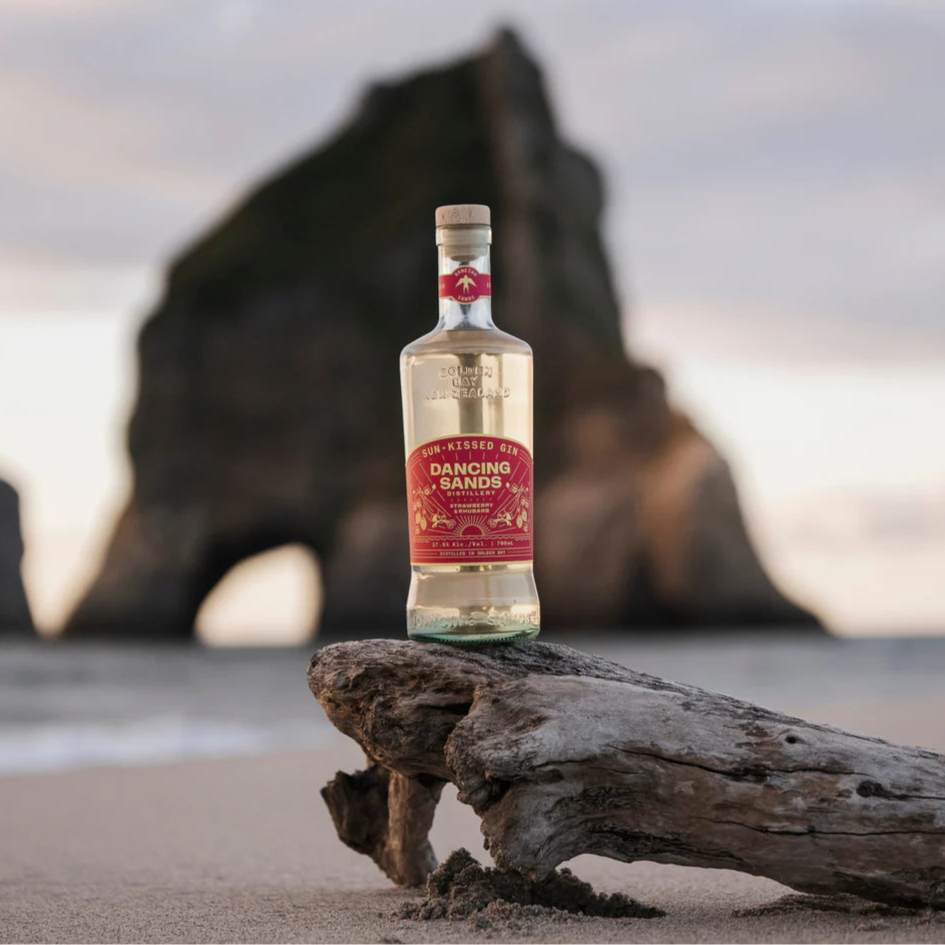 Load image into Gallery viewer, Bottle of 'Dancing Sands Sun Kissed Gin' on a piece of driftwood with a beach and rock formation in the background